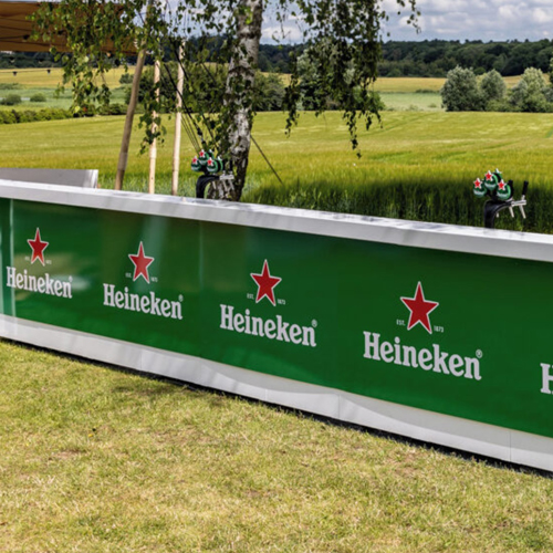 Outdoor grassy field with a green Heineken banner featuring red stars along a low advertising barrier.