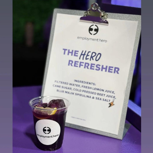 Purple hero refresher drink in a cup with a lemon wedge, placed beside a clipboard sign that reads 'THE HERO REFRESHER' from Employment Hero.