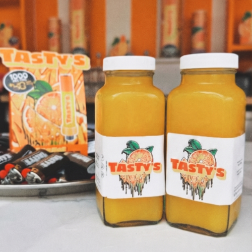 Two orange juice bottles with a white cap and a TASTY'S label on a counter, with orange snack packages in the background.