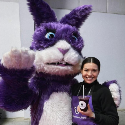 Smiling woman posing with a large purple bunny mascot waving at the camera; she holds a purple drink cup in front of her.