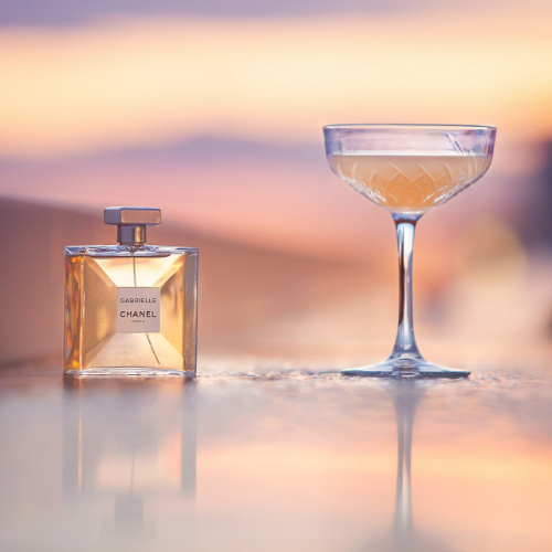 Chanel Gabrielle perfume bottle and a coupe glass on a reflective surface at sunset.