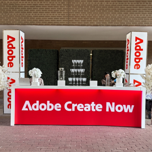 Promotional booth for Adobe Create Now with a bright red front panel reading 'Adobe Create Now', flanked by white pillars bearing red Adobe logos and floral arrangements. Behind the counter are glasses and people.]