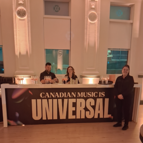 Three staff behind a reception bar with a banner reading 'CANADIAN MUSIC IS UNIVERSAL' in a bright, elegant room.