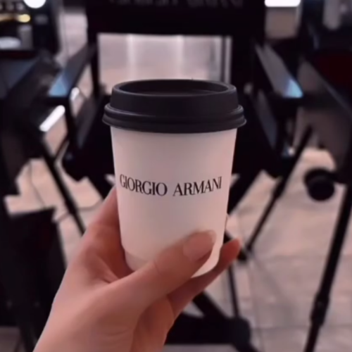 Hand holding a white disposable coffee cup with a black lid displaying the Giorgio Armani logo in a store setting.
