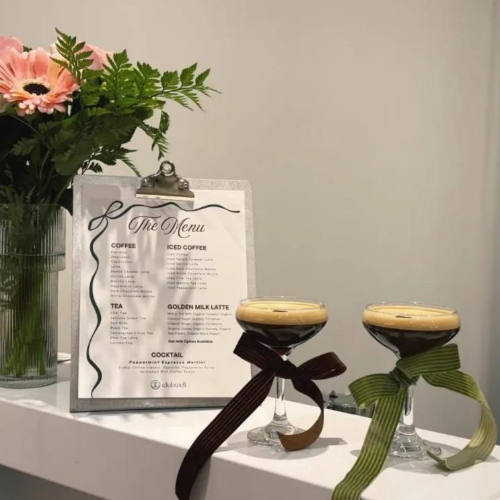 Clipboard menu on a white counter beside a vase of pink flowers, with two stemmed coffee drinks tied with ribbons.
