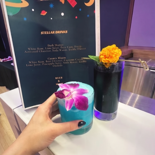 Hand holding a bright blue cocktail with a purple orchid garnish in front of a menu board and a vase with a yellow flower on the counter.