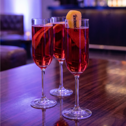 Three red cocktails in tall fluted glasses with citrus twists on a polished bar, under bluish lounge lighting in the background.
