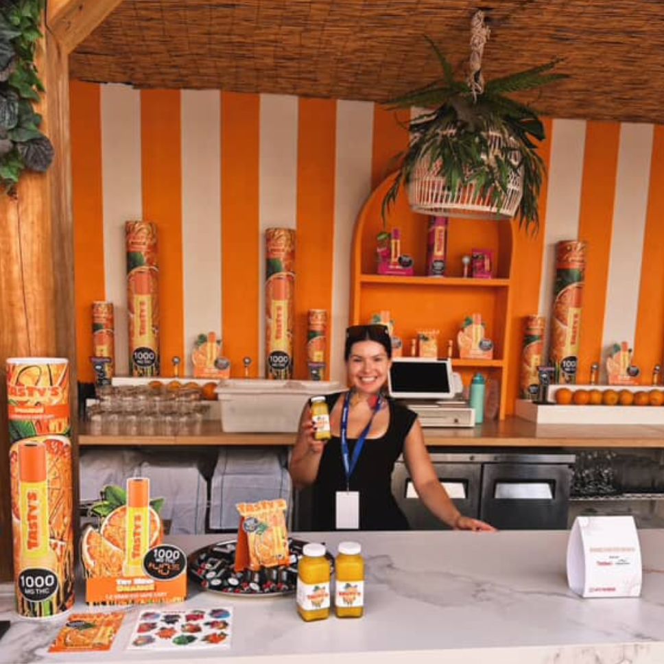 Smiling woman behind a bright orange-striped juice bar with bottled drinks and snacks on display.