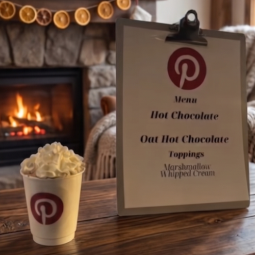 Cup of whipped-cream‑topped hot chocolate with a Pinterest logo beside a clipboard menu in a cozy room with a lit fireplace.