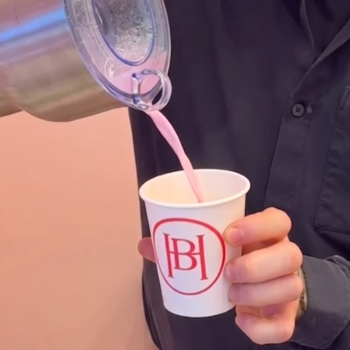 Person pours pink beverage from a pitcher into a white cup with a red circular logo.