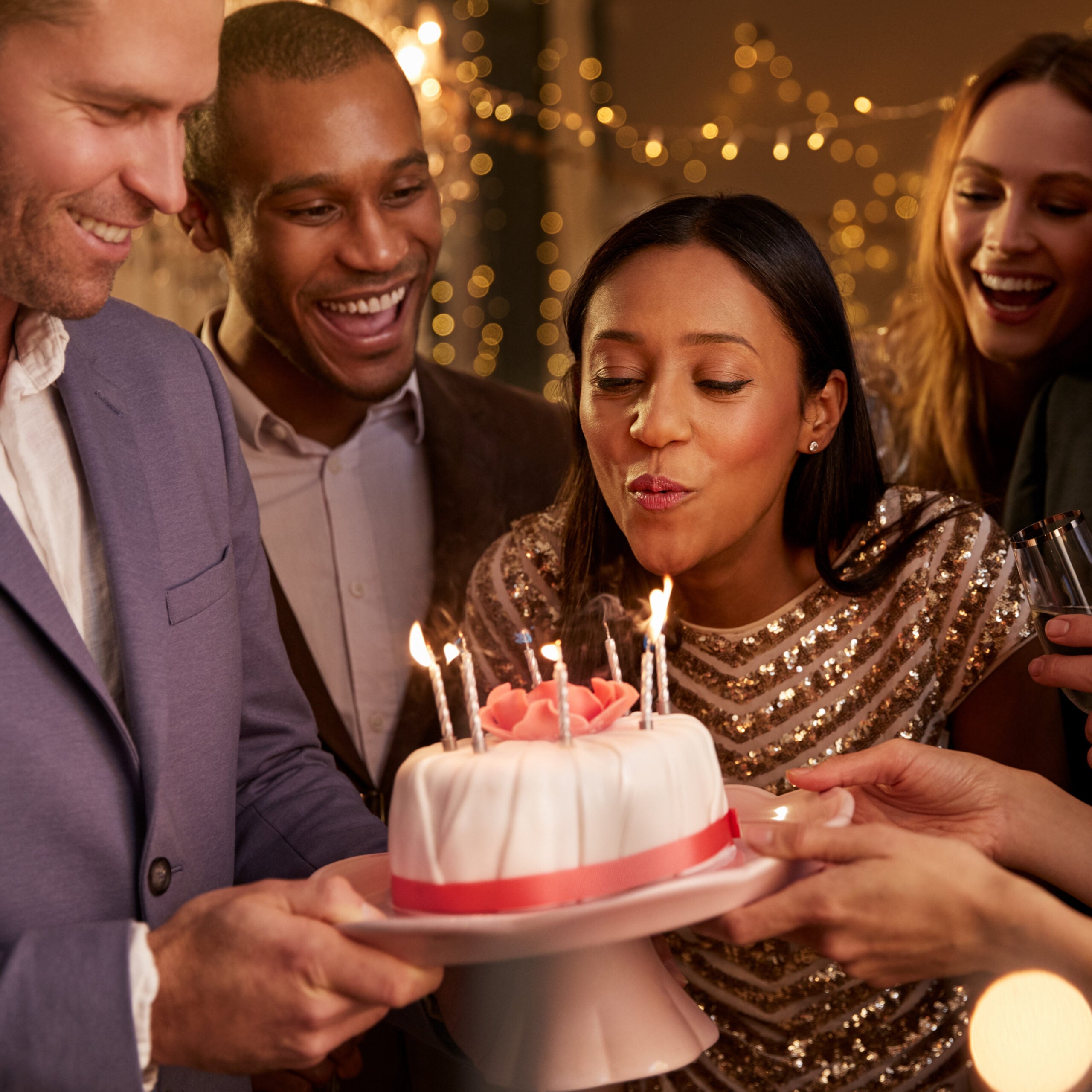 Friends gathered around a birthday cake with lit candles, smiling and celebrating in a warm, festive setting.