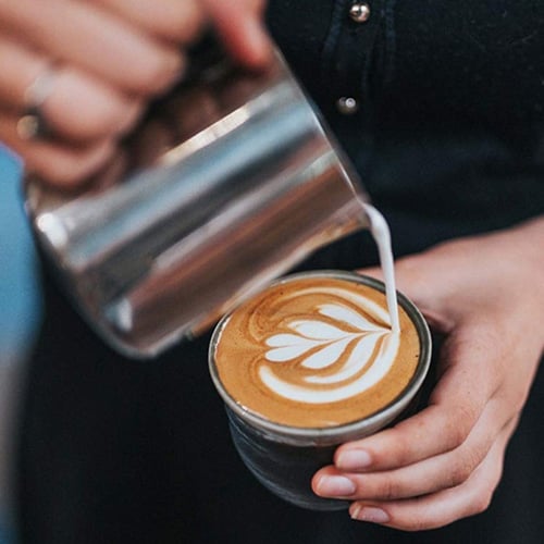 Barista pouring steamed milk to create latte art in a cup of coffee small metal pitcher in hand