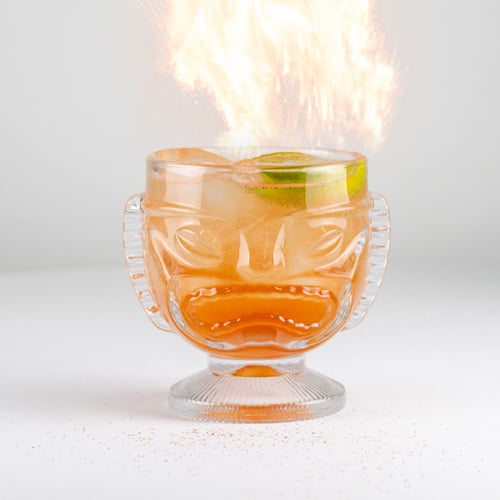 Flaming tiki glass with a face-shaped mug, orange cocktail, lime wedge, and a flame shooting from the top against a white background.