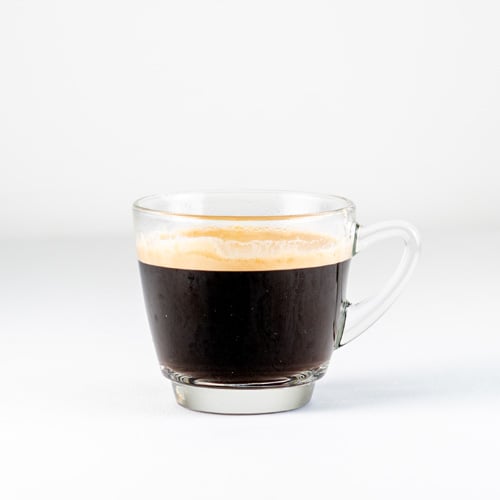 Clear glass cup of black coffee with a creamy crema on top on a white surface.
