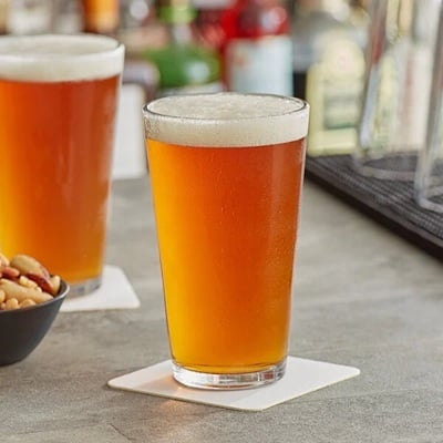 Two pint glasses of amber beer with foamy heads on white coasters at a bar, blurred bottles in the background
